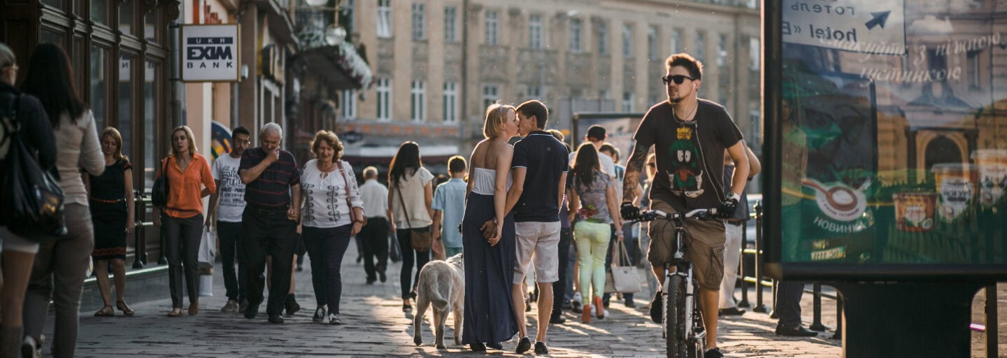 Homem em bicicleta na rua duma cidade do norte da Europa, com outras pessoas em fundo, incluindo um casal que se beija