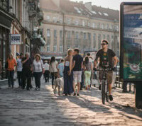 Homem em bicicleta na rua duma cidade do norte da Europa, com outras pessoas em fundo, incluindo um casal que se beija
