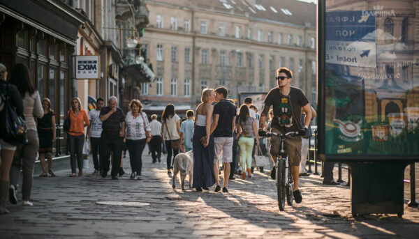 Homem em bicicleta na rua duma cidade do norte da Europa, com outras pessoas em fundo, incluindo um casal que se beija