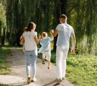 mãe e pai passeiam num parque solarengo enquanto seguram pelas mãos um filho que salta