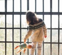 Criança virada para uma janela pela qual entra luz, de costas para nós, segura uma boneca-bebé pelo braço