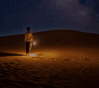 homem caminha no deserto, sob um céu estrelado, levando um luzeiro na mão