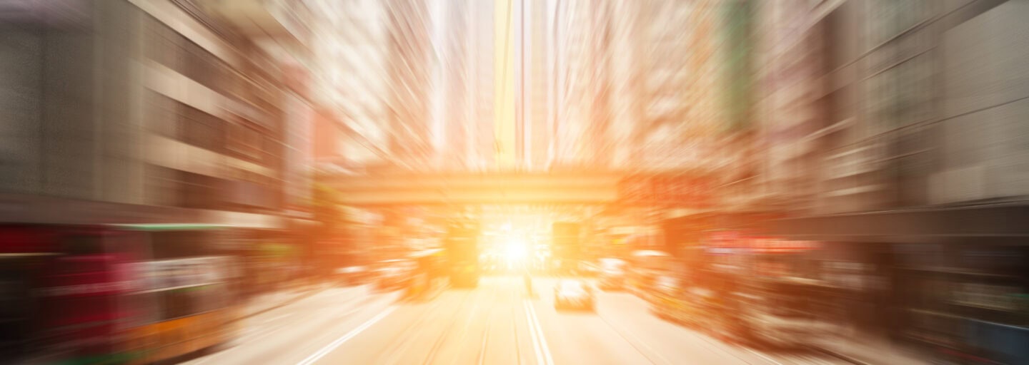 rua ladeada de prédios altos, com luz de pôr do sol ao fundo, distorcida pela velocidade de quem se move