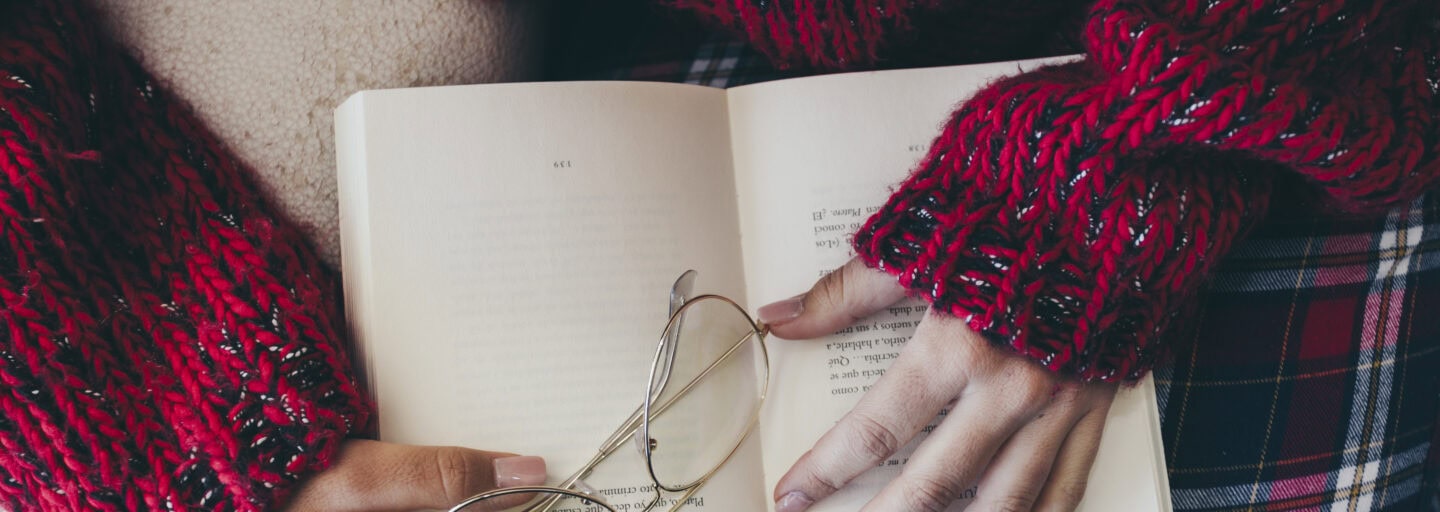 mulher vista de cima, com livro sobre o colo, onde repousam os seus óculos e as suas mãos
