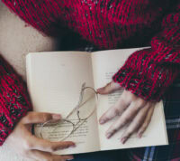 mulher vista de cima, com livro sobre o colo, onde repousam os seus óculos e as suas mãos