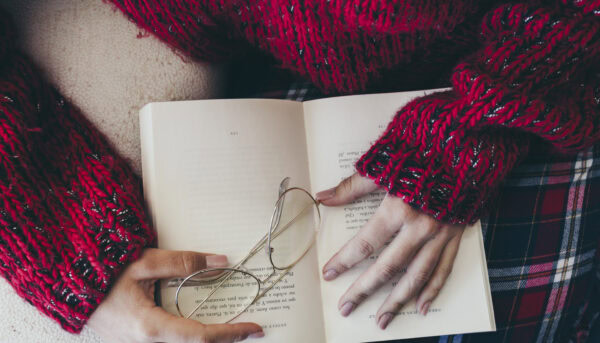 mulher vista de cima, com livro sobre o colo, onde repousam os seus óculos e as suas mãos