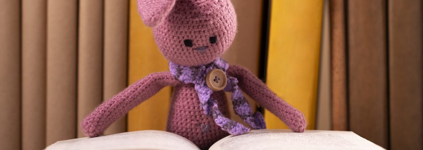 coelho rosa em croché, com cachecol rosa e um botão a fazer de alfinete de peito, debruça-se sobre livro aberto, com livros indistintos como fundo