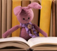 coelho rosa em croché, com cachecol rosa e um botão a fazer de alfinete de peito, debruça-se sobre livro aberto, com livros indistintos como fundo