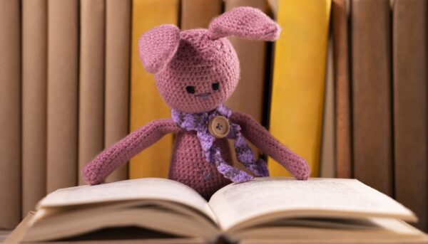 coelho rosa em croché, com cachecol rosa e um botão a fazer de alfinete de peito, debruça-se sobre livro aberto, com livros indistintos como fundo