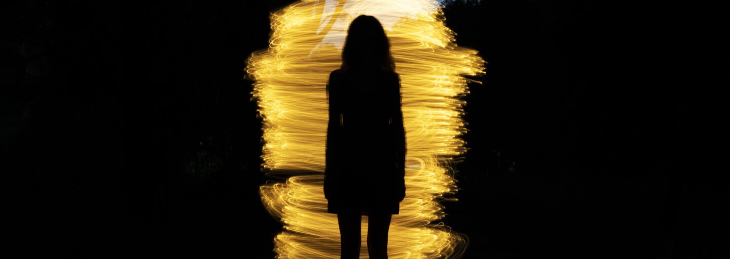imagem escura, em que se denota um fundo de árvores e uma espiral de várias luzes a formar um casulo, diante do qual se nota a silhueta duma mulher