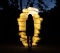 imagem escura, em que se denota um fundo de árvores e uma espiral de várias luzes a formar um casulo, diante do qual se nota a silhueta duma mulher
