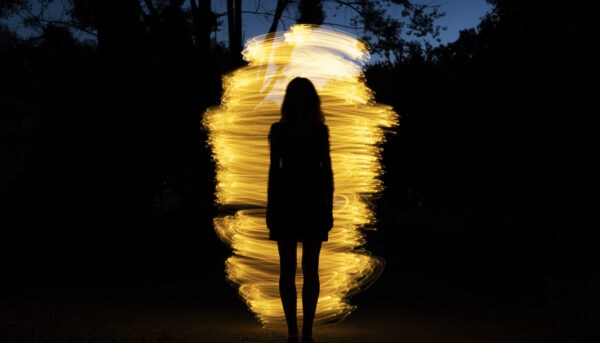 imagem escura, em que se denota um fundo de árvores e uma espiral de várias luzes a formar um casulo, diante do qual se nota a silhueta duma mulher