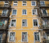 foto vertical de um edifício antigo, de cor amarela, com algumas janelas partidas