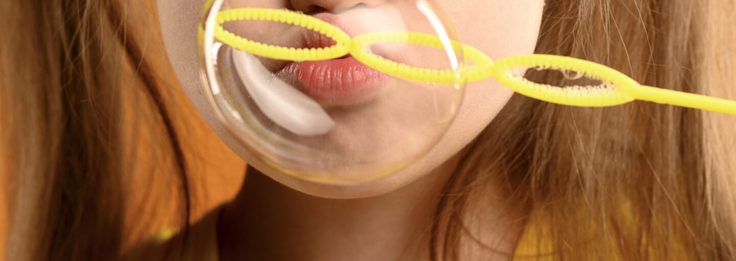 criança ruiva, em fundo laranja e vestida de amarelo, sopra uma bola de sabão