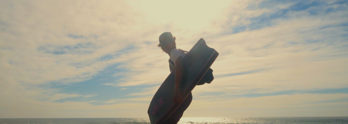Silhueta de bodyboarder que se vira para a câmara enquanto caminha para o mar, num momento de luz difusa