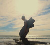 Silhueta de bodyboarder que se vira para a câmara enquanto caminha para o mar, num momento de luz difusa