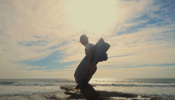 Silhueta de bodyboarder que se vira para a câmara enquanto caminha para o mar, num momento de luz difusa