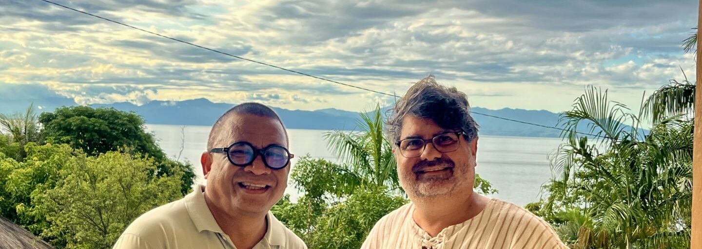 Dois jesuítas lado a lado, a sorrir para a câmara. Ambos usam óculos e camisas claras. Atrás deles vê‑se uma paisagem verde com palmeiras, um grande corpo de água e montanhas ao fundo, sob um céu parcialmente nublado