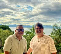 Dois jesuítas lado a lado, a sorrir para a câmara. Ambos usam óculos e camisas claras. Atrás deles vê‑se uma paisagem verde com palmeiras, um grande corpo de água e montanhas ao fundo, sob um céu parcialmente nublado