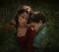 Duas pessoas sentam‑se juntas numa floresta verde e luminosa. Uma veste vermelho, a outra verde. Estão encostadas, de olhos fechados, num momento de calma e intimidade, envolvidas pela luz suave que atravessa as folhas.
