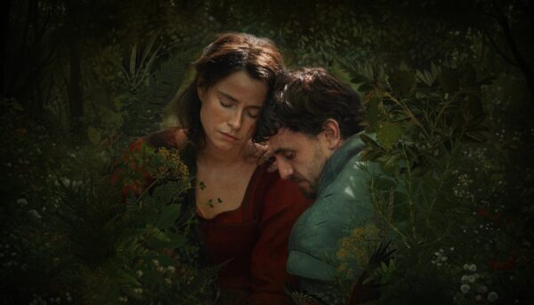 Duas pessoas sentam‑se juntas numa floresta verde e luminosa. Uma veste vermelho, a outra verde. Estão encostadas, de olhos fechados, num momento de calma e intimidade, envolvidas pela luz suave que atravessa as folhas.