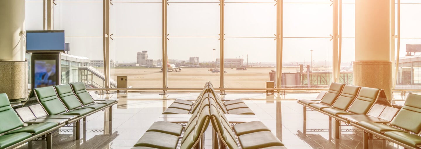 uma fila de cadeiras verdes estão em primeiro plano, com um fundo em vidro transparente donde se vê, entre luz ofuscante de pôr do sol, uma pista de aeroporto