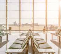 uma fila de cadeiras verdes estão em primeiro plano, com um fundo em vidro transparente donde se vê, entre luz ofuscante de pôr do sol, uma pista de aeroporto