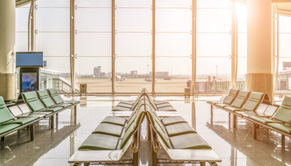 uma fila de cadeiras verdes estão em primeiro plano, com um fundo em vidro transparente donde se vê, entre luz ofuscante de pôr do sol, uma pista de aeroporto