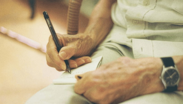 “Mãos do P. Roque Cabral, sj, enquanto escreve num pequeno bloco de notas, segurando uma caneta. Usa camisa clara e um relógio de pulso. O fundo está desfocado, destacando apenas o gesto de escrita.