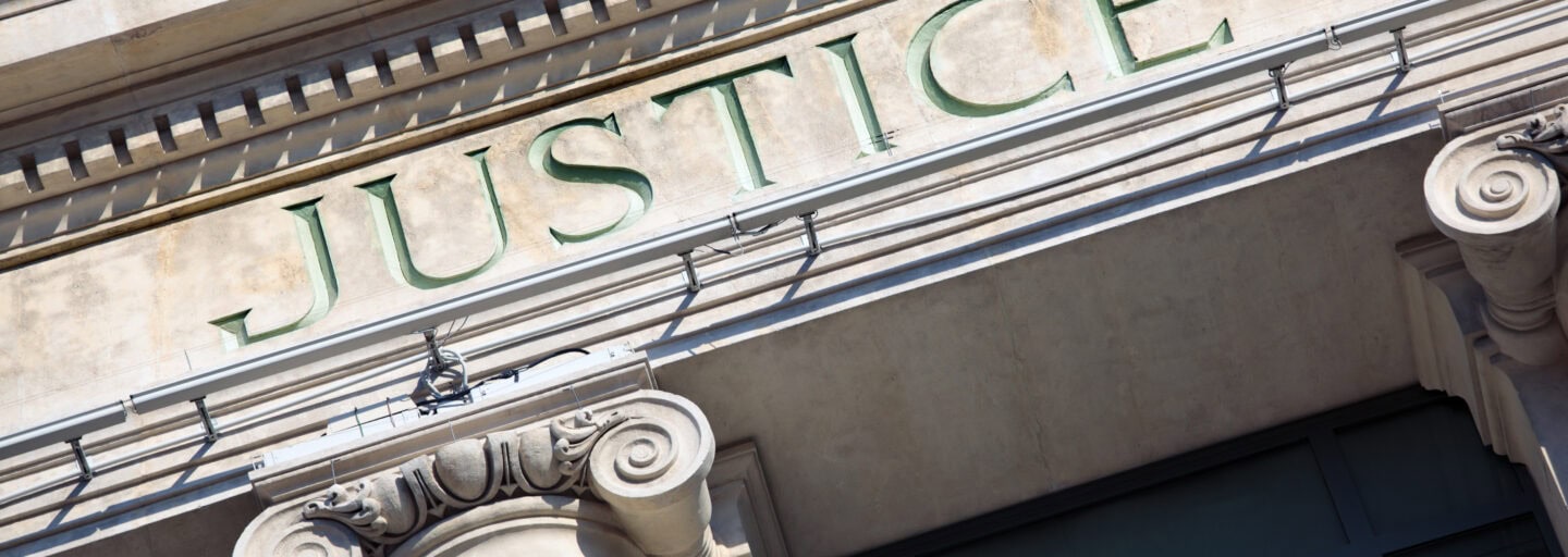 fachada de um edifício clássico, com grandes colunas de pedra. Acima delas, está gravada a palavra JUSTICE, pintada a verde, sugerindo tratar‑se de um tribunal ou instituição ligada à justiça.
