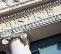 fachada de um edifício clássico, com grandes colunas de pedra. Acima delas, está gravada a palavra JUSTICE, pintada a verde, sugerindo tratar‑se de um tribunal ou instituição ligada à justiça.