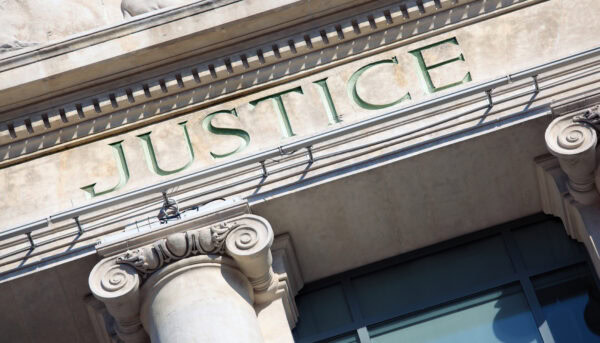 fachada de um edifício clássico, com grandes colunas de pedra. Acima delas, está gravada a palavra JUSTICE, pintada a verde, sugerindo tratar‑se de um tribunal ou instituição ligada à justiça.